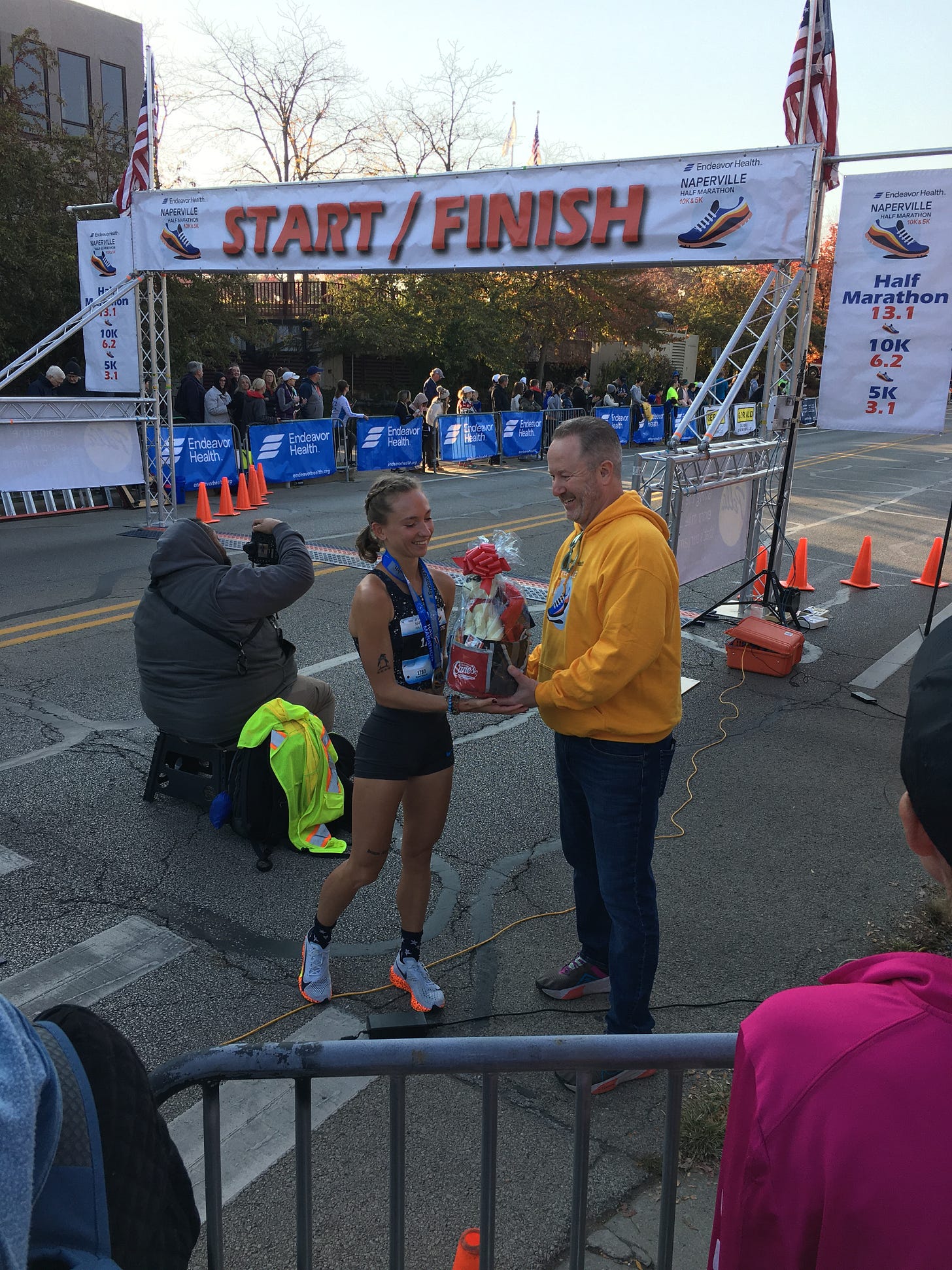 Fast woman runner recieves prize for winning the half marathon. The start line/ finish line of the road race is in the background. This was during the awards ceremony of an outdoor event.