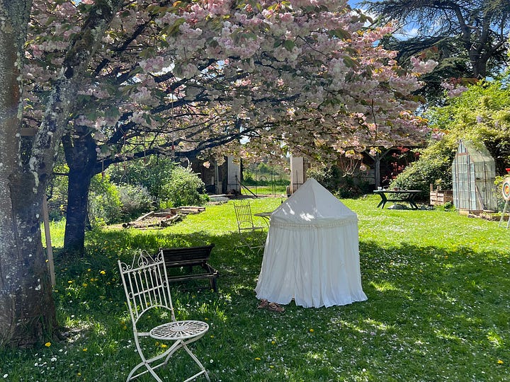 Four photos of a walled French garden in spring: a canopy tent on the grass under blossoming trees, a wooden playhouse and swing set beneath a cherry tree in full pink bloom, a young girl planting seedlings in a raised bed beside a wheelbarrow, and a trampoline framed by climbing roses and a ribbon archway.