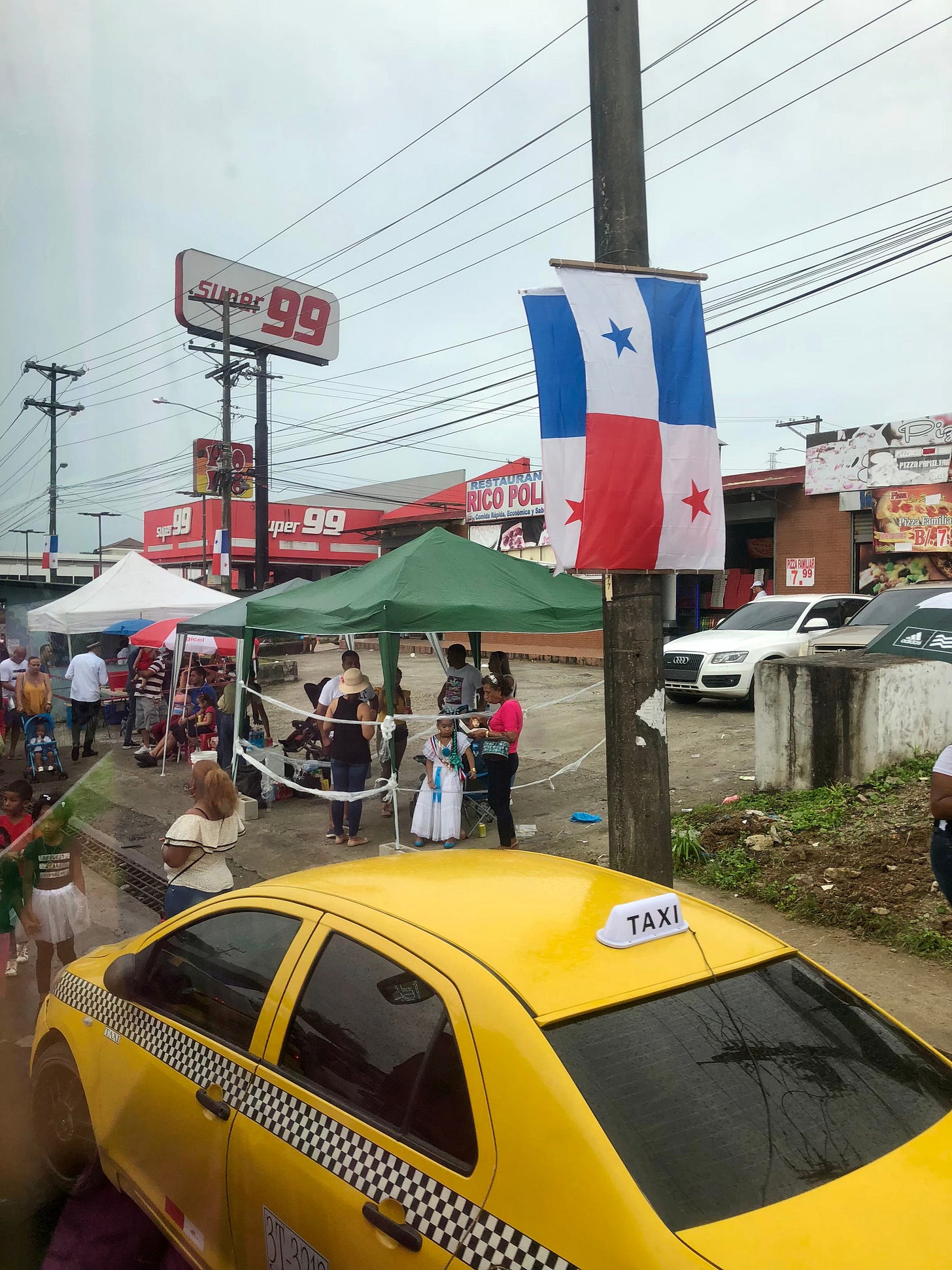 Image from the streets of Panama, featuring a yellow taxi, stalls with umbrellas for shade, a few stores, and people strolling about