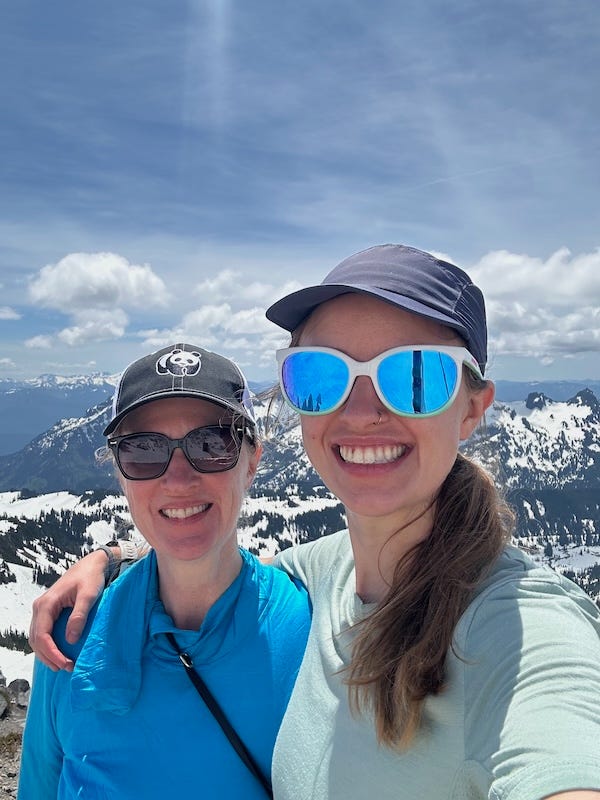 Julie Carrick Dalton and her adult daughter standing on a mountaintop. Both are smiling and wearing sunglasses and baseball caps. A blue sky and snow-covered peaks in the background.