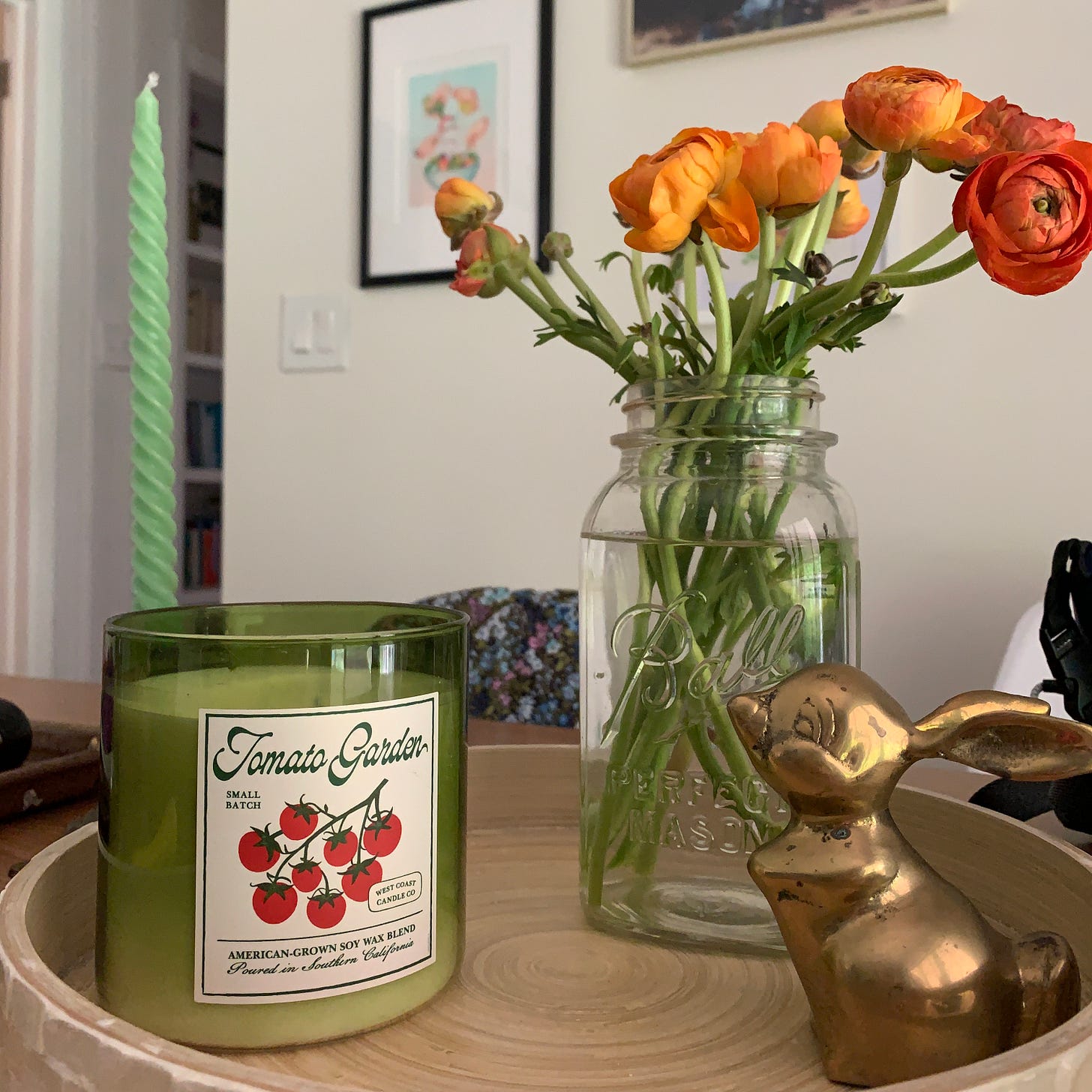 A centerpiece arrangement on a kitchen table of a large, green candle called "Tomato Garden," orange ranunculus in a mason jar, and a vintage MCM brass rabbit with long ears.