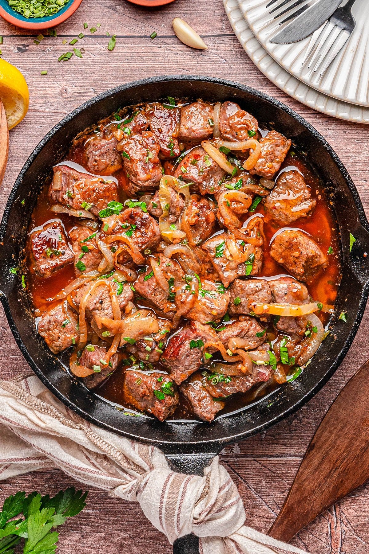 A cast iron skillet filled with cooked beef cubes, sliced onions, and chopped herbs in a brown sauce, placed on a wooden table with plates and utensils nearby.