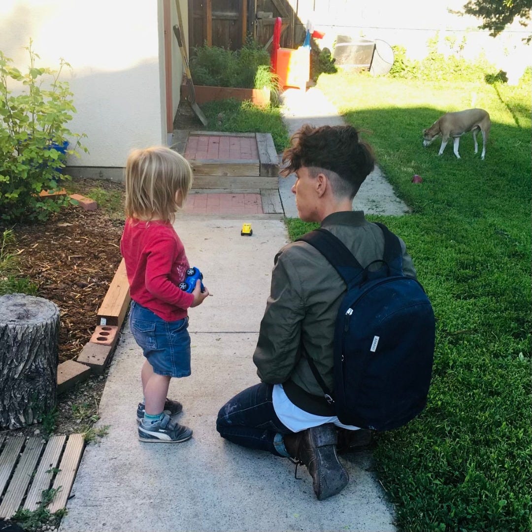 Andrea looking at their godson in a back yard. Their godson is holding a toy truck and looking at a dog sniffing the grass. Andrea looking at their godson in a back yard. Their godson is holding a toy truck and looking at a dog sniffing the grass.