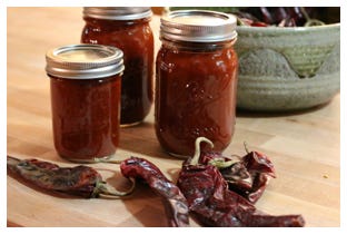 Picture of three mason jars of red jam and some red chillies in front of them.