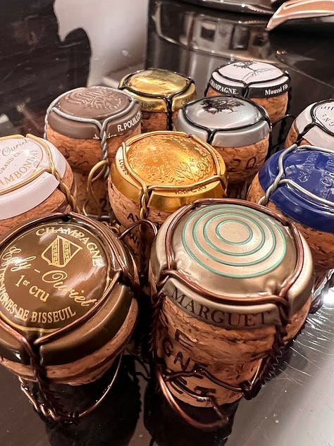 A gallery of cork closures: a natural cork branded 'Vila Voltaire' resting on a wine barrel table, a cork being pulled from a bottle with a waiter's corkscrew, and a cluster of Champagne corks and cages from various producers
