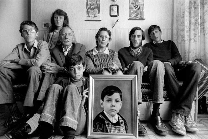 Una famiglia seduta su un divano in bianco e nero, composta da sette persone di diverse età. Al centro è posato un ritratto incorniciato di un ragazzo. La scena è in un ambiente domestico semplice con decorazioni alle pareti.