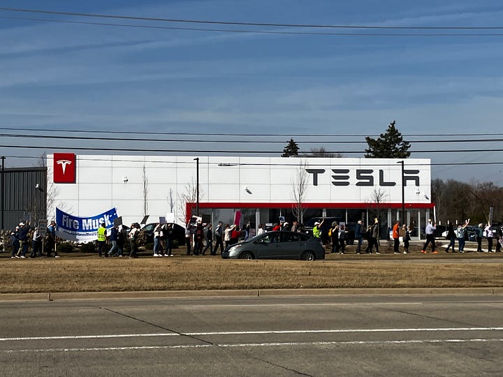 Protestors walk with picket signs in front of a Tesla dealership in Ann Arbor, Mich. The dealership is a long white building with the word Tesla. People with placards march in front of the building.