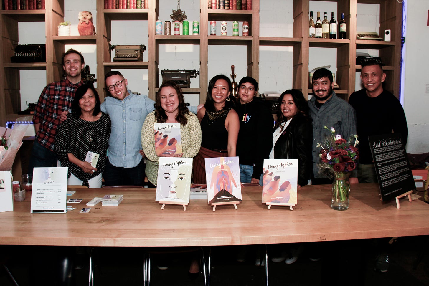 Group of 8 people all smiling and standing behind a table with displays of the Living Hyphen magazine.