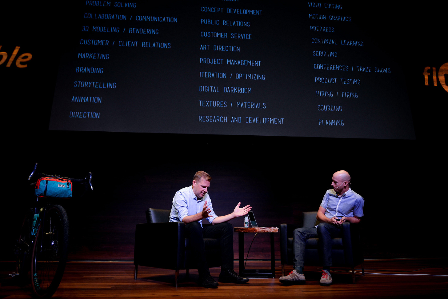 A stage scene featuring two men, I'm one of them, in casual attire seated in chairs, engaged in a conversation. Me gesturing animatedly while the other listens intently. A bicycle with a colorful handlebar bag stands to the left. In the background, a large screen displays a long list of terms including 'problem solving,' 'collaboration,' '3D modeling,' 'marketing,' 'project management,' 'video editing,' and 'planning,' set against a dark backdrop. A stage scene featuring two men, I'm one of them, in casual attire seated in chairs, engaged in a conversation. Me gesturing animatedly while the other listens intently. A bicycle with a colorful handlebar bag stands to the left. In the background, a large screen displays a long list of terms including 'problem solving,' 'collaboration,' '3D modeling,' 'marketing,' 'project management,' 'video editing,' and 'planning,' set against a dark backdrop.
