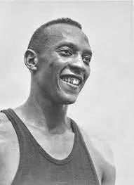 Black and white photograph of American track and field athlete Jesse Owens mid-race at the 1936 Berlin Olympics, smiling with visible joy.