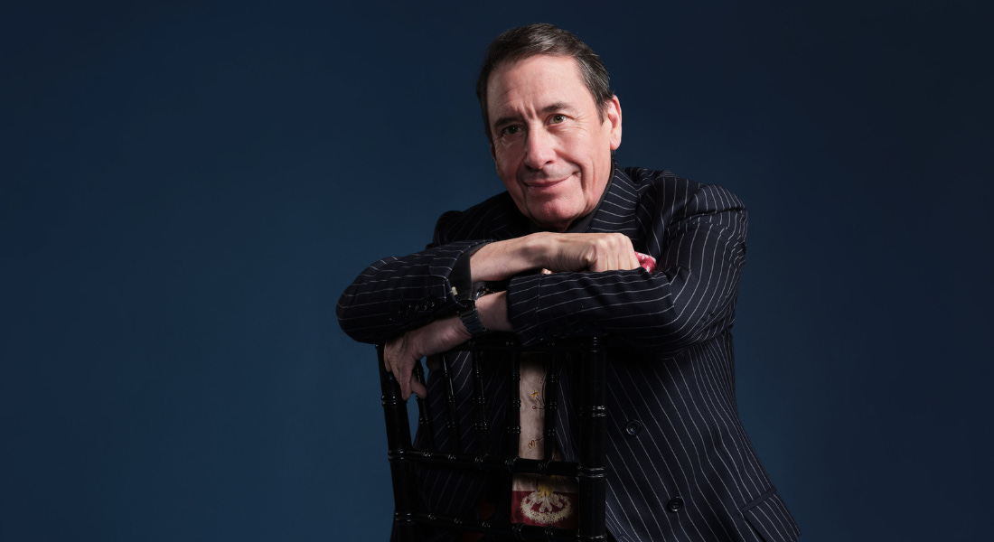 Jools Holland - a man wearing a pin-striped suit with black hair - sitting backwards on a chair, on a dark blue background Jools Holland - a man wearing a pin-striped suit with black hair - sitting backwards on a chair, on a dark blue background