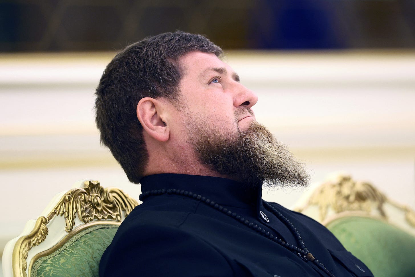 Ramzan Kadyrov on an ornate chair with a full, long beard looking slightly upwards with a contemplative expression. He’s wearing a dark-coloured shirt with a high collar. Around his neck is a string of black braver beads or a bead necklace