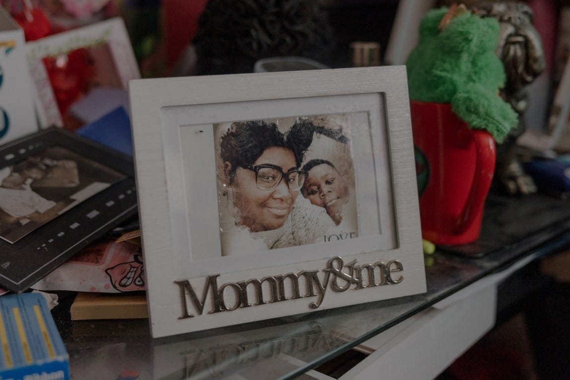 A white picture frame with the words “Mommy & me” sits on a glass desk surrounded by other family photos. The picture frame contains a photo of JJ resting his head on his mother’s shoulder as they both smile, looking at the camera.