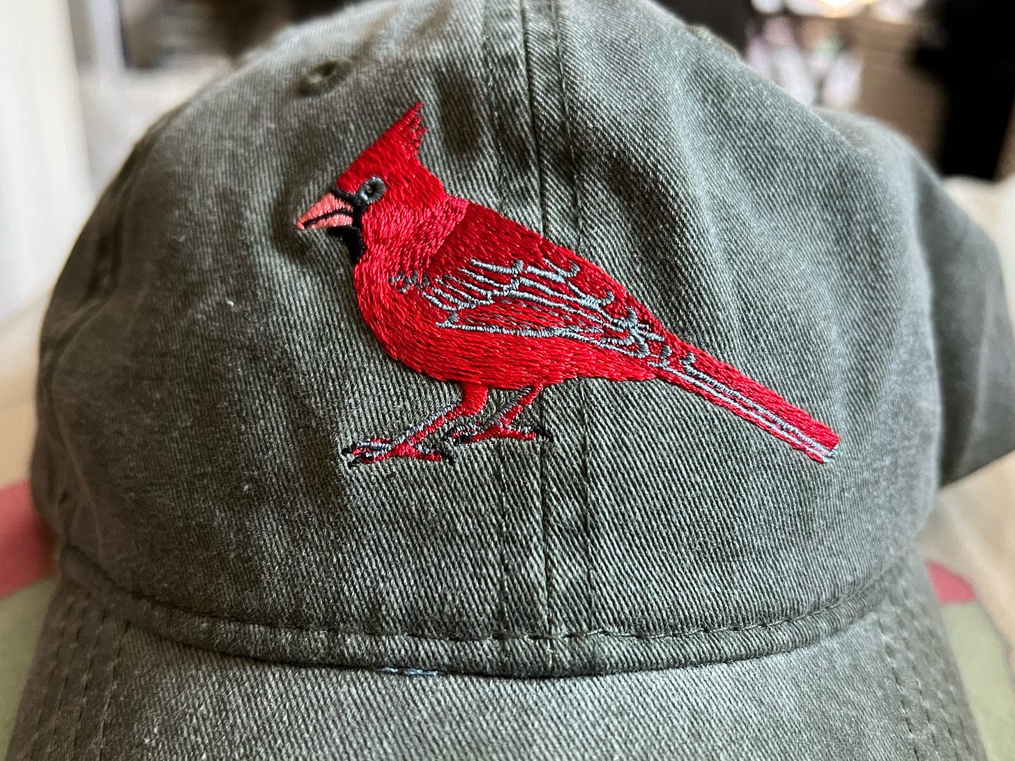 a forest green hat with a northern cardinal - red bird with a chunky orange bill - embroidered on the front, sitting on a bed a forest green hat with a northern cardinal - red bird with a chunky orange bill - embroidered on the front, sitting on a bed