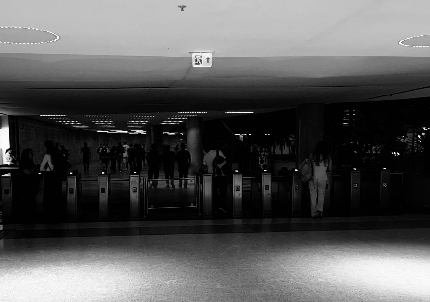 Foto preto e branca de catracas de um prédio comercial. No todo há um ícone que indica saída.