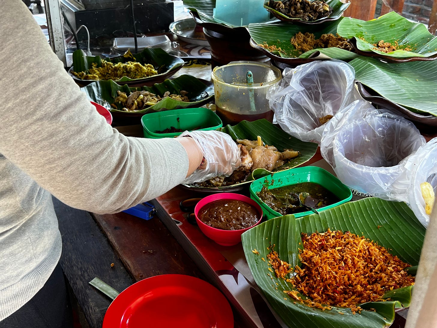 Behind a counter, banana-leaf trays of prepared dishes and a bowl of sambal, a gloved hand assembling a plate from them.