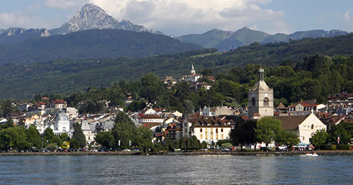 Lakefront town - Evian-les-Bains -  with colorful buildings and church tower, set against lush green hills and a mountain range under a partly cloudy sky.