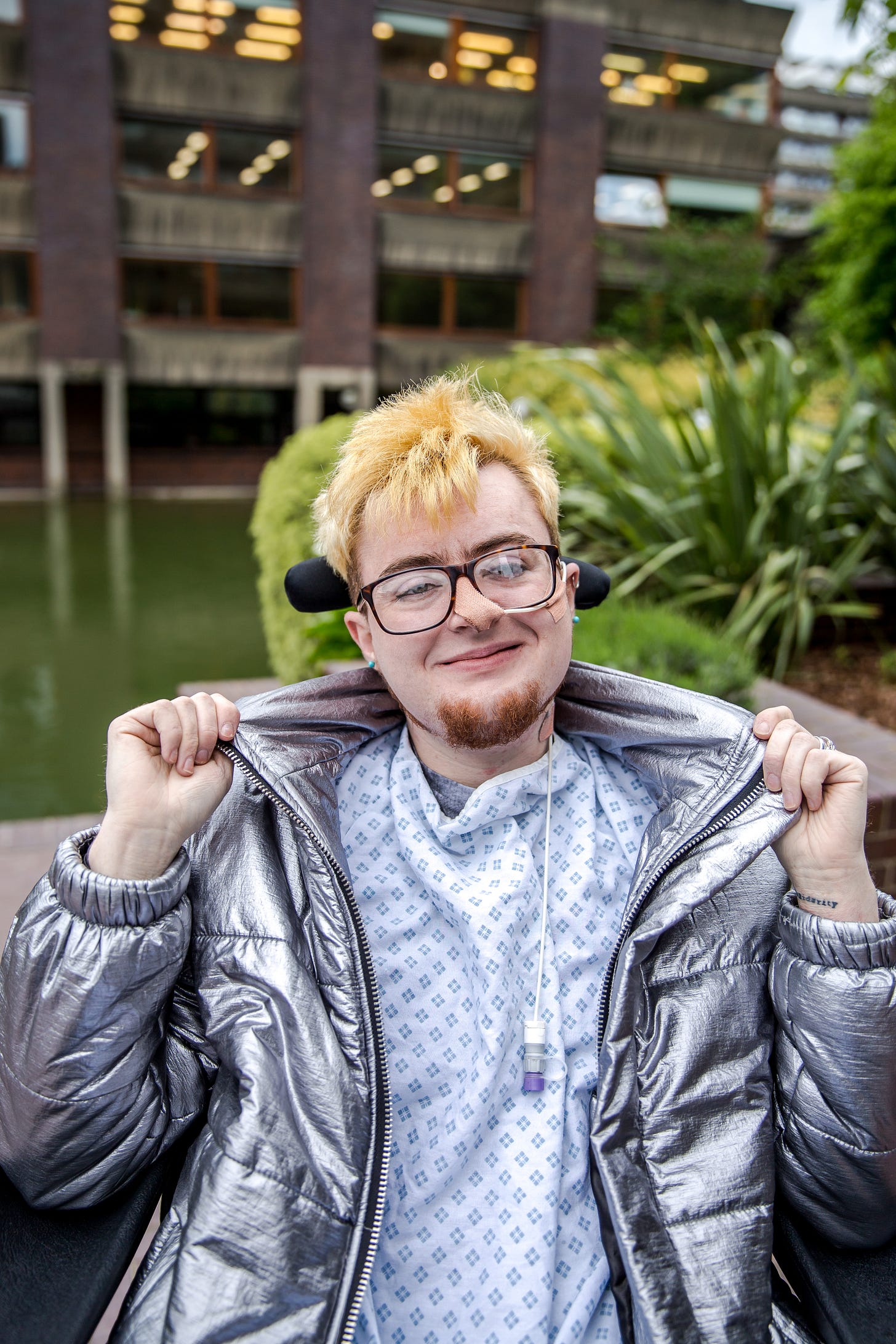 Jamie Hale, a white electric wheelchair user with a shock of blonde hair sits before the Barbican lake. There is a feeding tube in their nose and they wear a hospital gown under a silver jacket they are holding open proudly