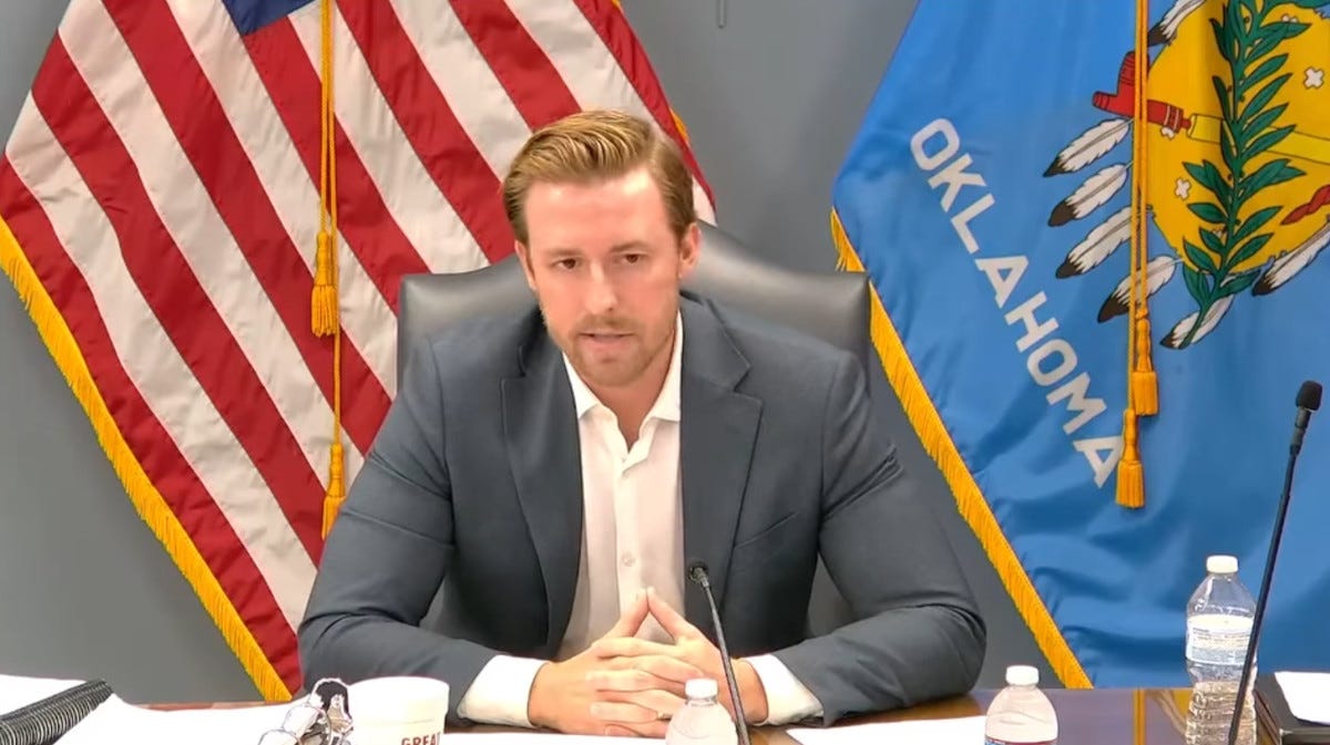 May speaking with U.S. and Oklahoma flags behind him.