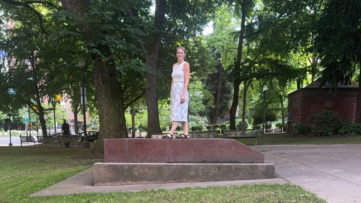 A middle-aged white woman in a white tanktop and skirt and with blond hair pulled back in a ponytail stands atop the empty base of a statue in Portland, Oregon. A middle-aged white woman in a white tanktop and skirt and with blond hair pulled back in a ponytail stands atop the empty base of a statue in Portland, Oregon.