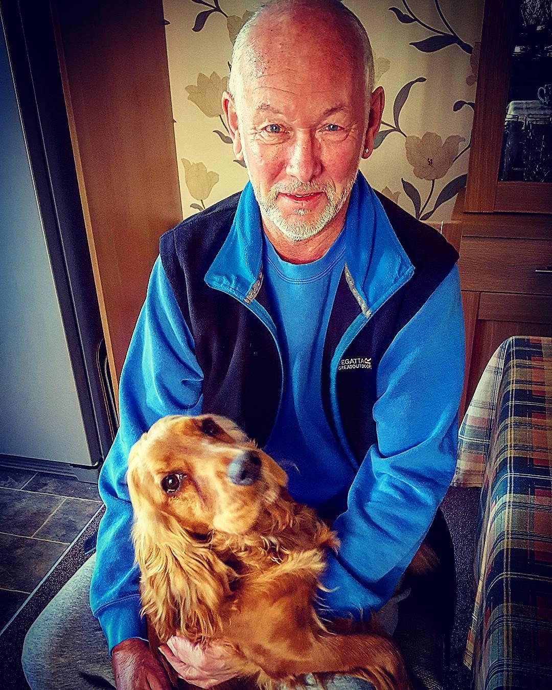 A smiling adult man with a shaved head and short grey beard is kneeling indoors, wearing a bright blue long-sleeved top and a dark sleeveless fleece. He is gently holding a golden-coloured Cocker Spaniel on his lap. The dog has long, wavy ears and looks up toward the man with a soft, relaxed expression. The setting appears to be a cosy home environment, with patterned wallpaper, a wooden cabinet, and a sofa visible in the background. The overall mood of the image is calm, affectionate, and warm, showing a close, trusting bond between the person and the dog.