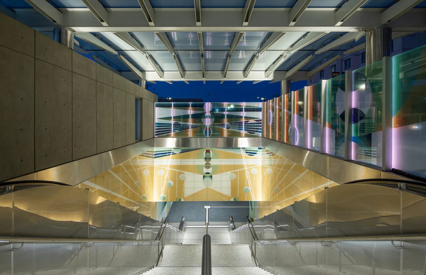 A wide view of a modern underground station entrance with glass railings and a large geometric artwork in blue, green, yellow, and purple integrated into the walls above the escalators. A wide view of a modern underground station entrance with glass railings and a large geometric artwork in blue, green, yellow, and purple integrated into the walls above the escalators.