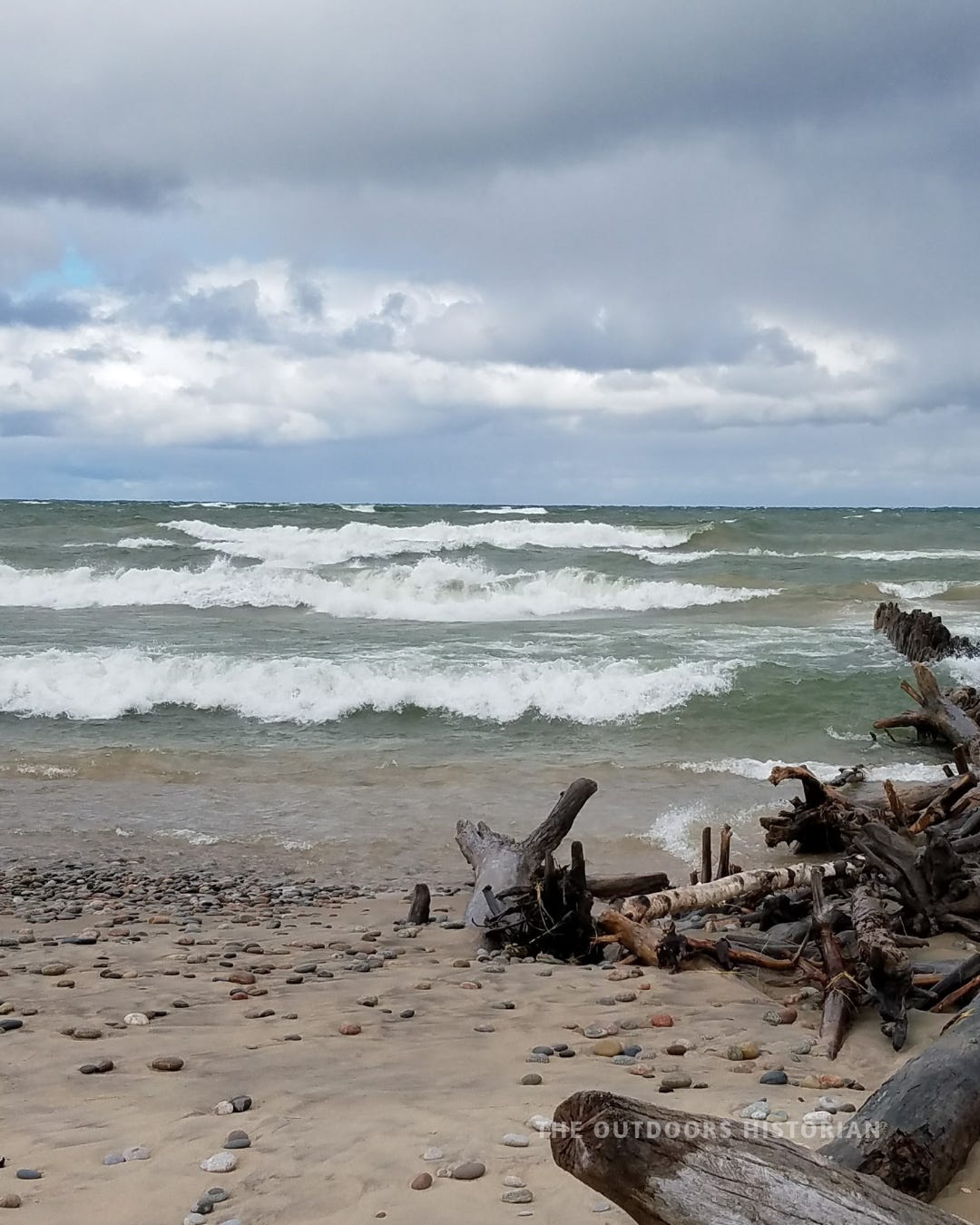 Whitecapped waves rolling onto a rocky Lake Superior beach under a heavy gray sky, with a large driftwood pile in the foreground.