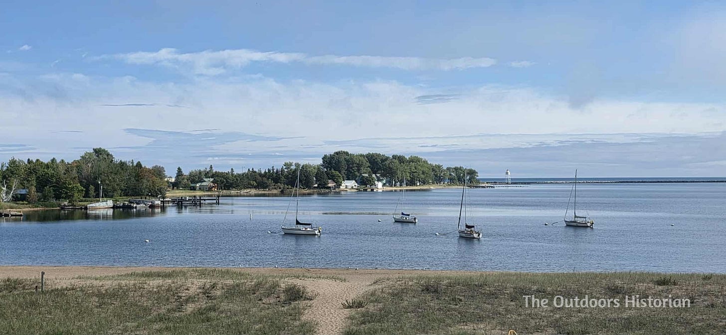 A view from a sand dune overlooking a beach and a harbor with four anchored sailboats. The lake is blue and the sky is blue with scattered white clouds.