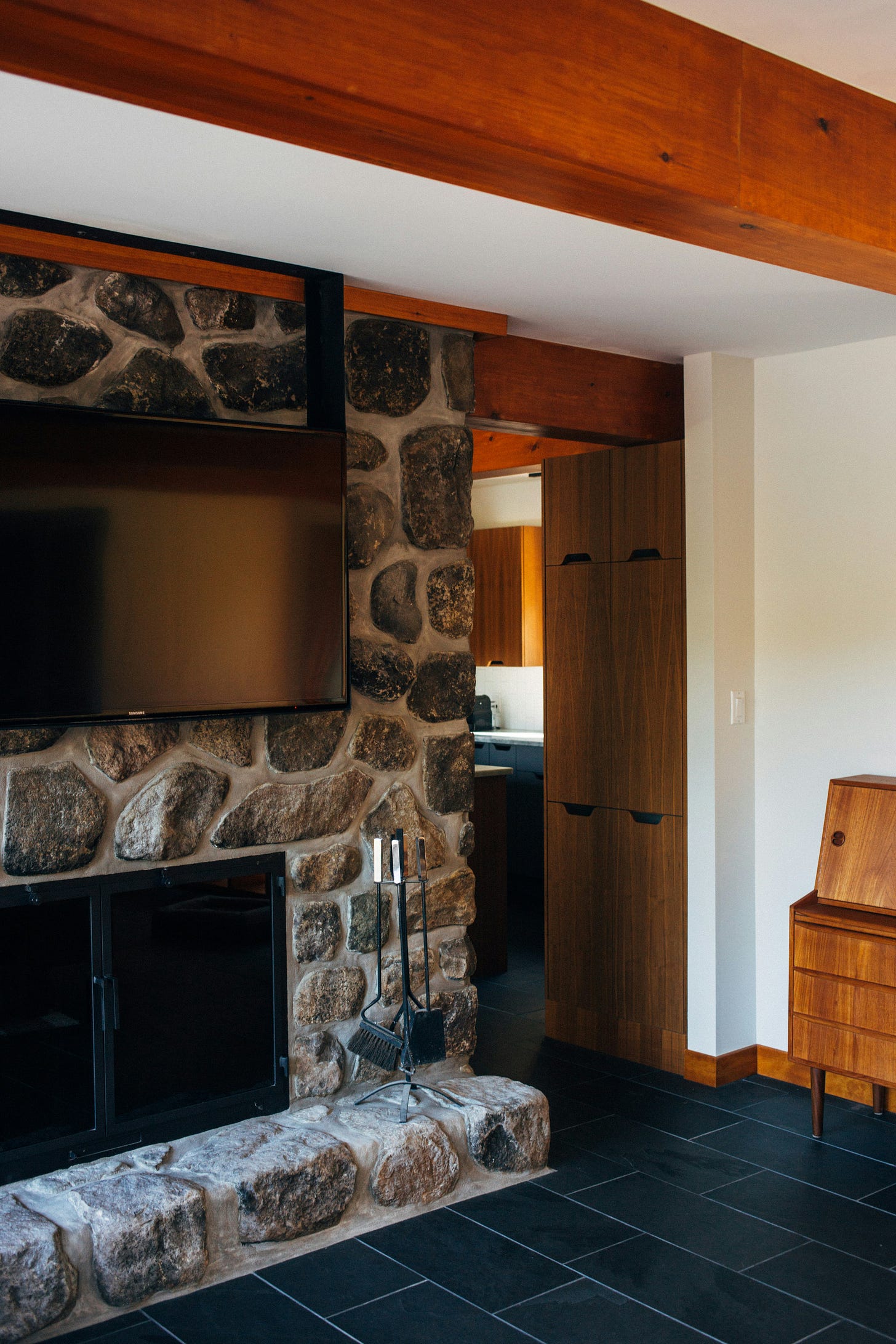 A fireplace made from many stones sits in the middle of a more modern, wooden walled house. The floor is black tile and the walls are painted white A fireplace made from many stones sits in the middle of a more modern, wooden walled house. The floor is black tile and the walls are painted white