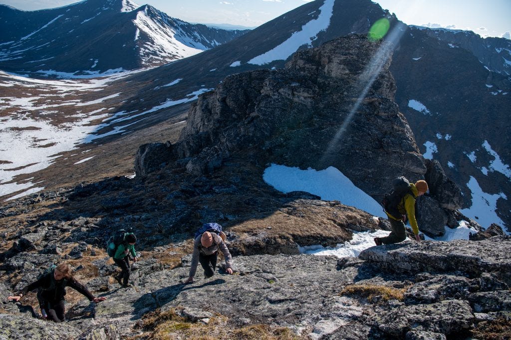 Four hikers scramble up a broad rib of sunlit granite along a high ridge, snow-streaked peaks and a deep valley behind them, a shaft of lens flare crossing the frame.