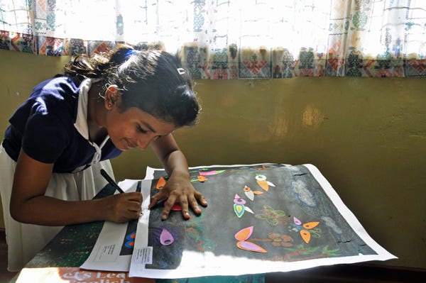 Young South Asian girl is wearing her blue and white uniform at school. She is bending over a piece of her colorful artwork, signing the bottom.