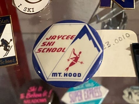 Left: A pinback button about 1" across sits in a museum display case. It was for the Jaycee Ski School and is red, white, and blue. Center: A sit-ski outrigger from the 1990s. It has an aluminum shaft and the very front tip of a ski attached to the end. Right: A white, black, and light blue button for the flying outriggers ski club. Their logo shows a pair of standing-ski outriggers, like crutches, with flags on them, waving in the wind..