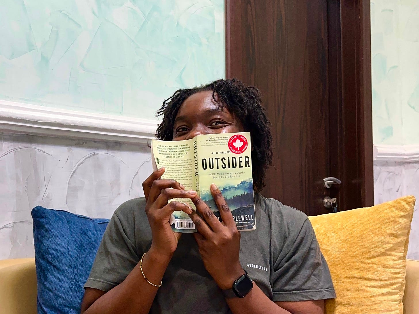 Folaa Oseni smiling while holding the book, "Outside" by Brett Popplewell. Folaa is wearing a grey tshirt while sitting on a blue and yellow couch. 