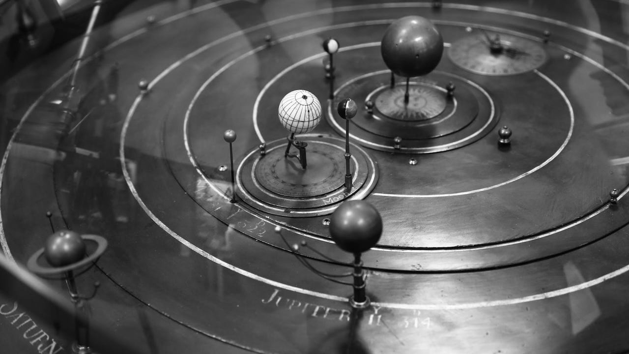 A monochrome close-up of a mechanical orrery model showing planets on metal arms orbiting a central sphere, with labeled circular tracks and smaller spherical bodies representing their moons.