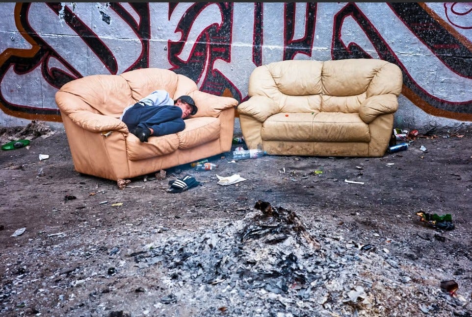 Two abandoned leather sofas and burnt debris on Turbo Island, Stokes Croft, Bristol — a man sleeping rough on one sofa beside a graffitied wall, at the site central to the 2026 village green inquiry. Two abandoned leather sofas and burnt debris on Turbo Island, Stokes Croft, Bristol — a man sleeping rough on one sofa beside a graffitied wall, at the site central to the 2026 village green inquiry.