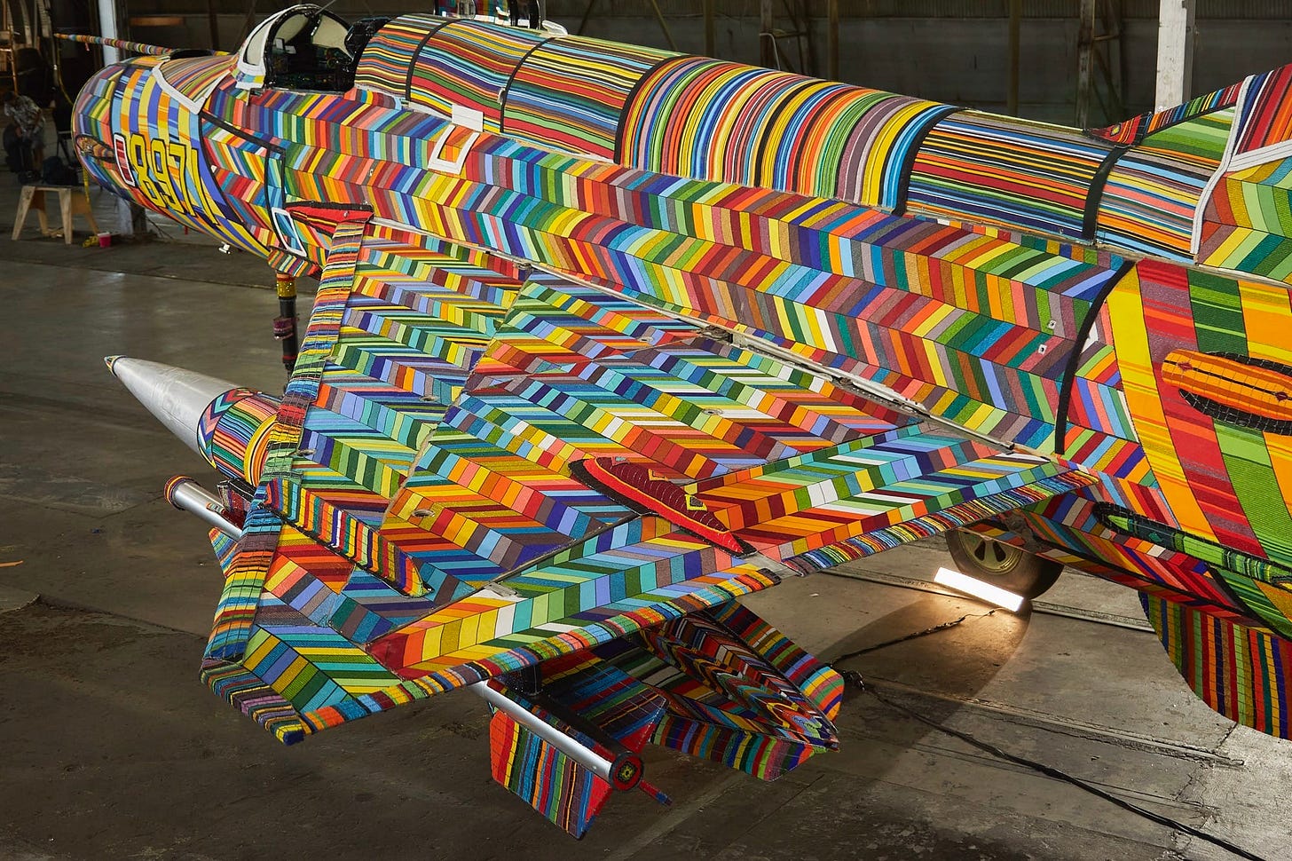 an overview of one wing and side of a fighter jet coated in colorful beaded patterns, sitting inside a hangar or warehouse