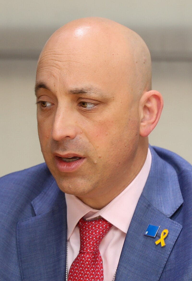 headshot, three quarters; bald man speaking, wearing red white spotted tie; yellow hostage support lapel pin
