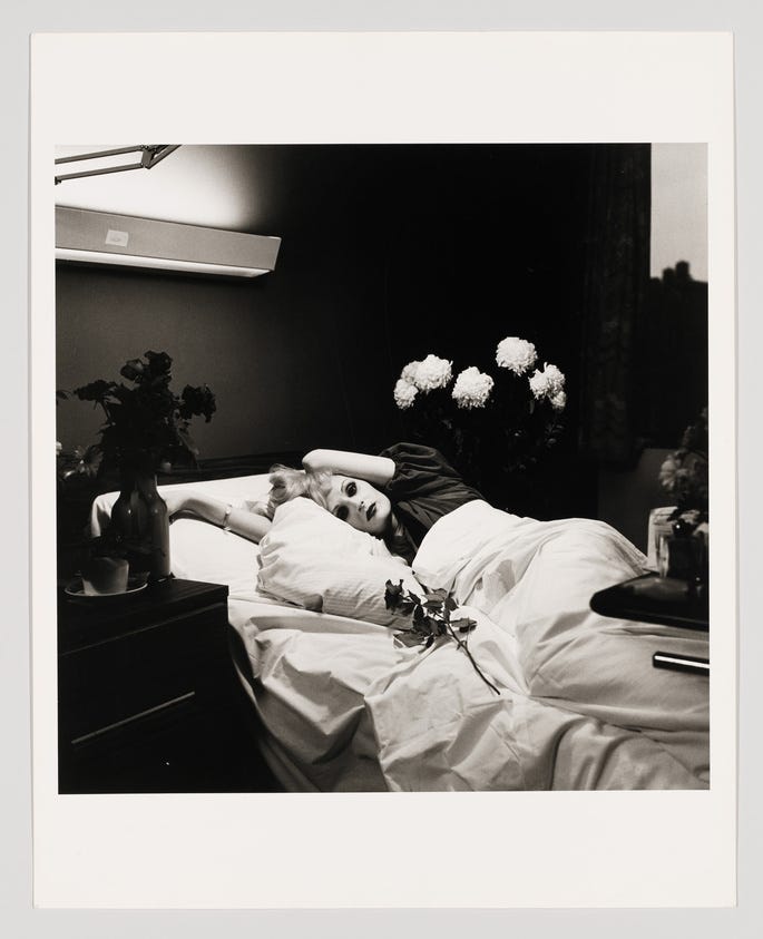 A black and white photograph depicting a woman lying on her back in bed, with her head resting on her arm. She appears to be gazing directly at the camera with an intense expression. The bed is adorned with white linens, and there are flowers in vases on the nightstands beside her. The room is dimly lit, creating a dramatic and moody atmosphere.