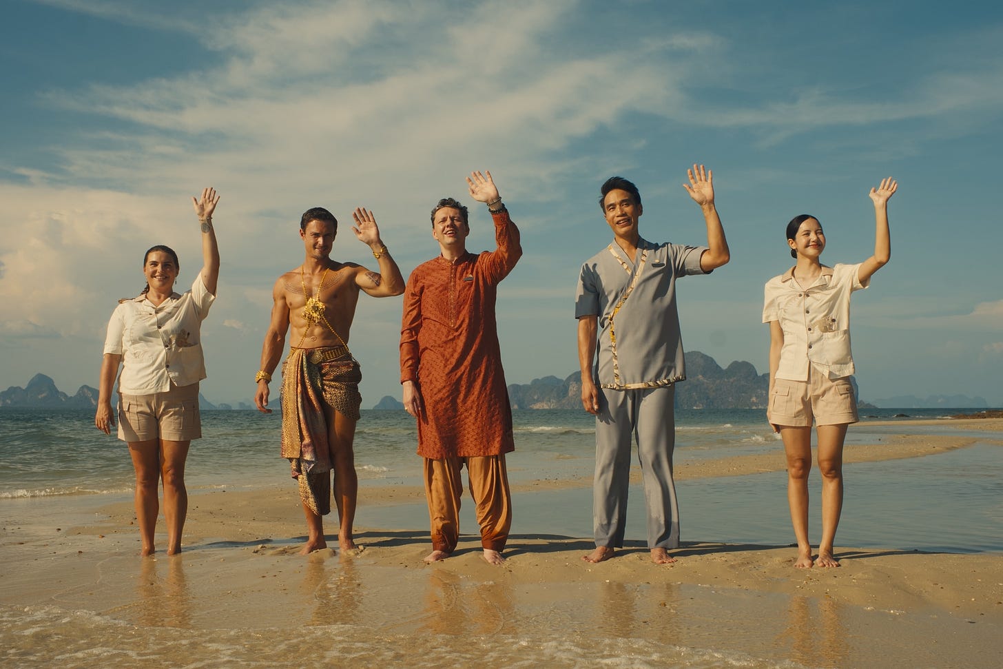 Morgana O'Reilly, Arnas Fedaravičius, Christian Friedel, Dom Hetrakul, and Lalisa Manobal as the staff of The White Lotus resort in Thailand, in a production still from Season 3 of HBO’s “The White Lotus.” They are standing by the sea and smiling and waving theatrically. Morgana O'Reilly, Arnas Fedaravičius, Christian Friedel, Dom Hetrakul, and Lalisa Manobal as the staff of The White Lotus resort in Thailand, in a production still from Season 3 of HBO’s “The White Lotus.” They are standing by the sea and smiling and waving theatrically.
