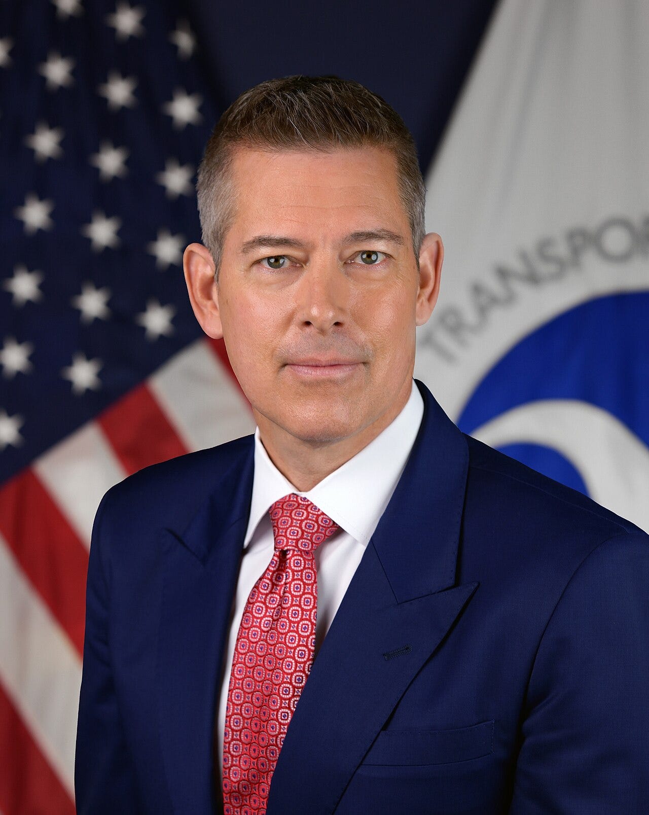 Official portrait of Sean Duffy; white man with dark hair greying on sides; blue suit white shirt red patterned tie; American flag and transportation department flag partially visible behind him.