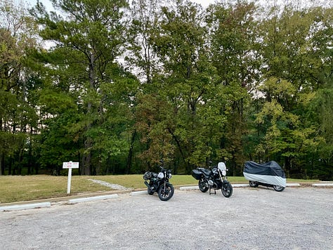 a road with trees, sign for tent sites B18, B19, B20, 3 motorcycles parked in the gravel near the tent sign