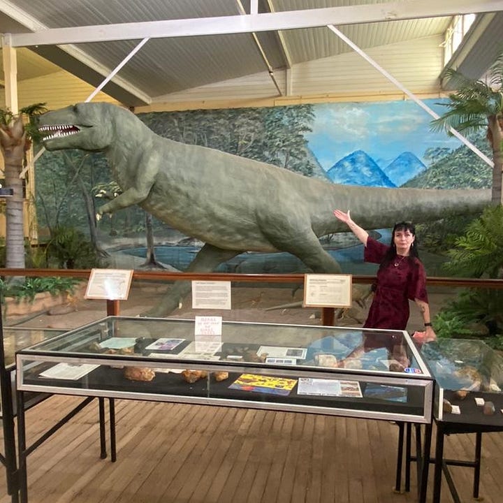 The first image is of a crowded bookstore, the walls are painted a dark green, and the shelves in the foreground and background are overflowing with books. The vibe is eclectic and welcoming. In the second image Kim Wilkins stands with her arm raised showing off a large replica dinosaur that she is standing beside. In the foreground is a display cabinet with books open on display.