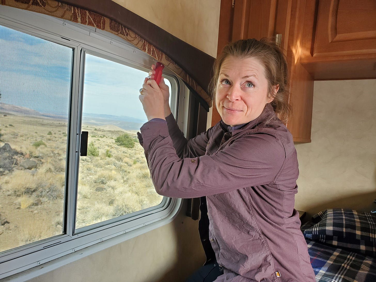 Charlotte inside the travel trailer looking up above the camera while she unscrews a brown window valance. 
