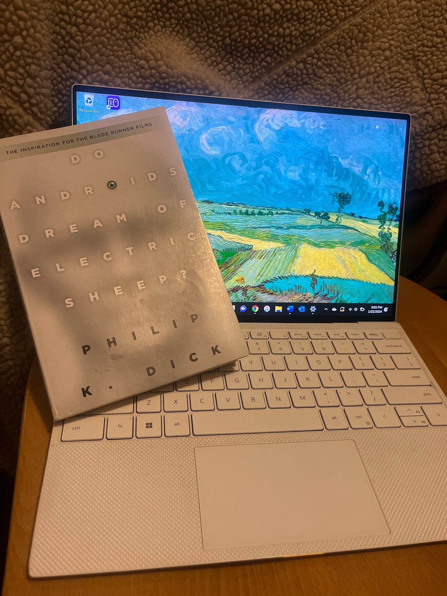 A copy of Philip K. Dick's novel Do Androids Dream of Electric Sheep, resting against the home screen of a laptop, which features an impressionist painting of a field beneath a blue sky. A copy of Philip K. Dick's novel Do Androids Dream of Electric Sheep, resting against the home screen of a laptop, which features an impressionist painting of a field beneath a blue sky.