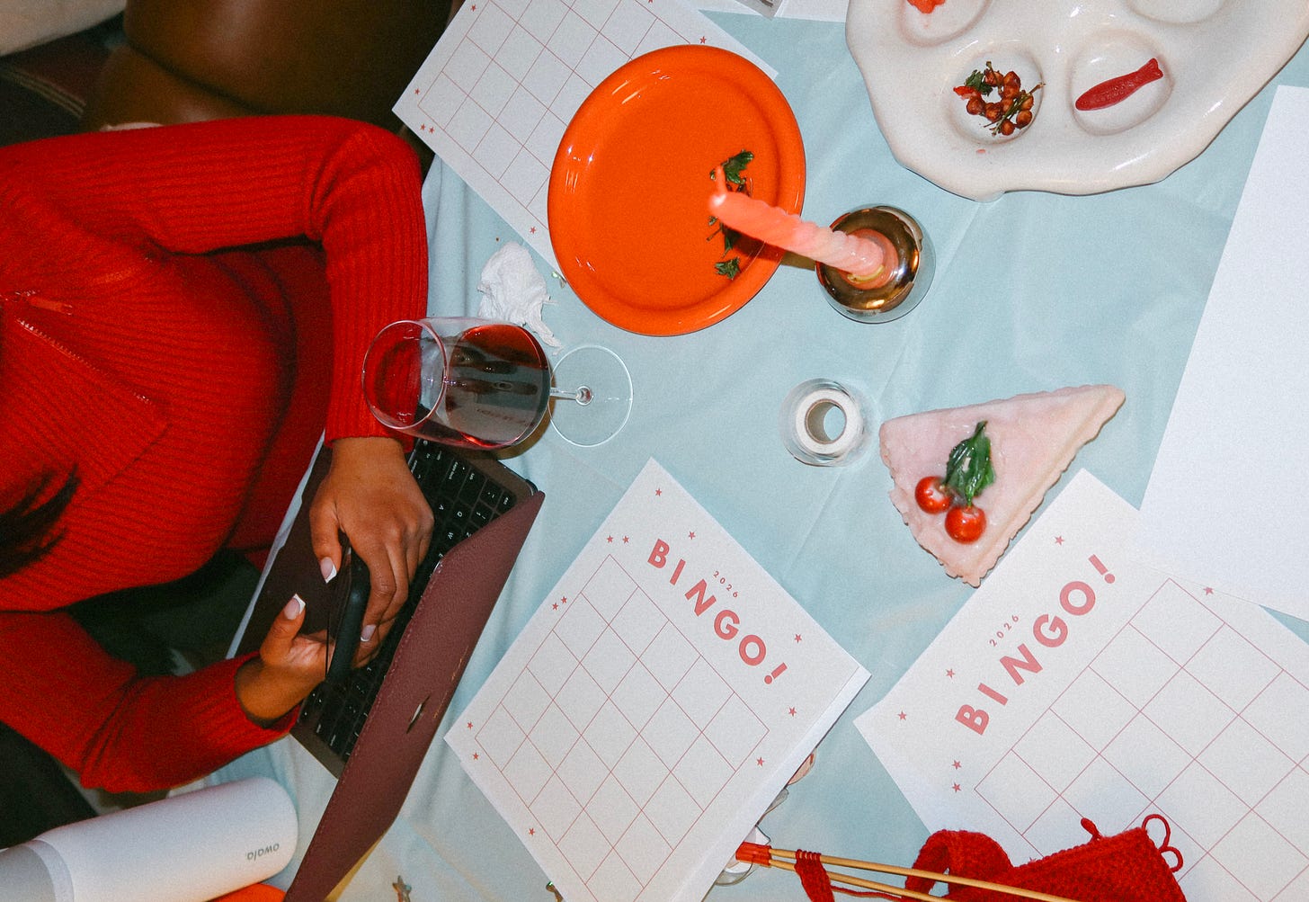 Person in a red sweater using a laptop at a table set with bingo cards, candles, and snacks
