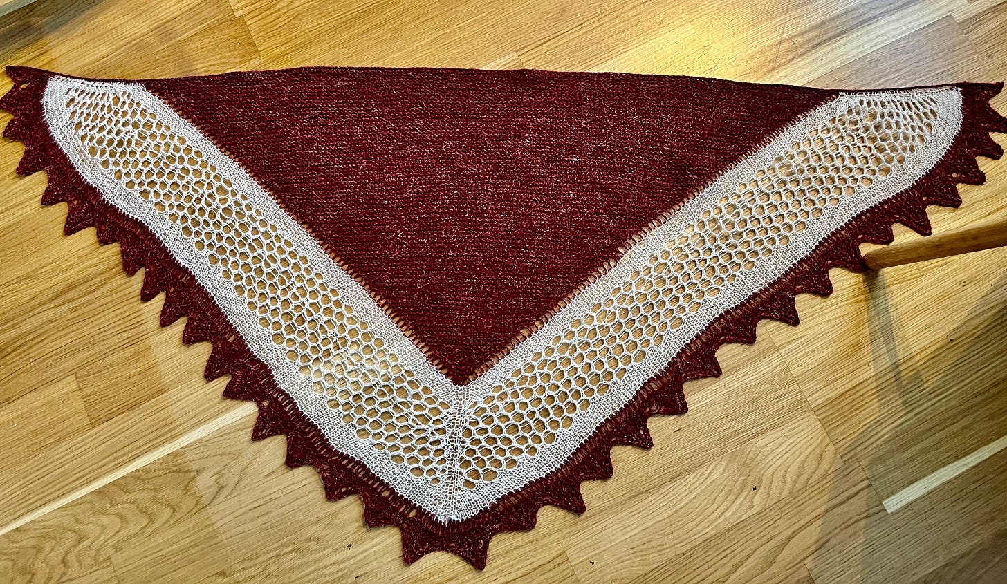 A finished triangular Shetland hap shawl, with a plain garter-stitch centre, a lace border in ecru wool, a contrasting colour panel of Bird's Eye lace, and a knitted-on edging in a wool-linen blend. A finished triangular Shetland hap shawl, with a plain garter-stitch centre, a lace border in ecru wool, a contrasting colour panel of Bird's Eye lace, and a knitted-on edging in a wool-linen blend.