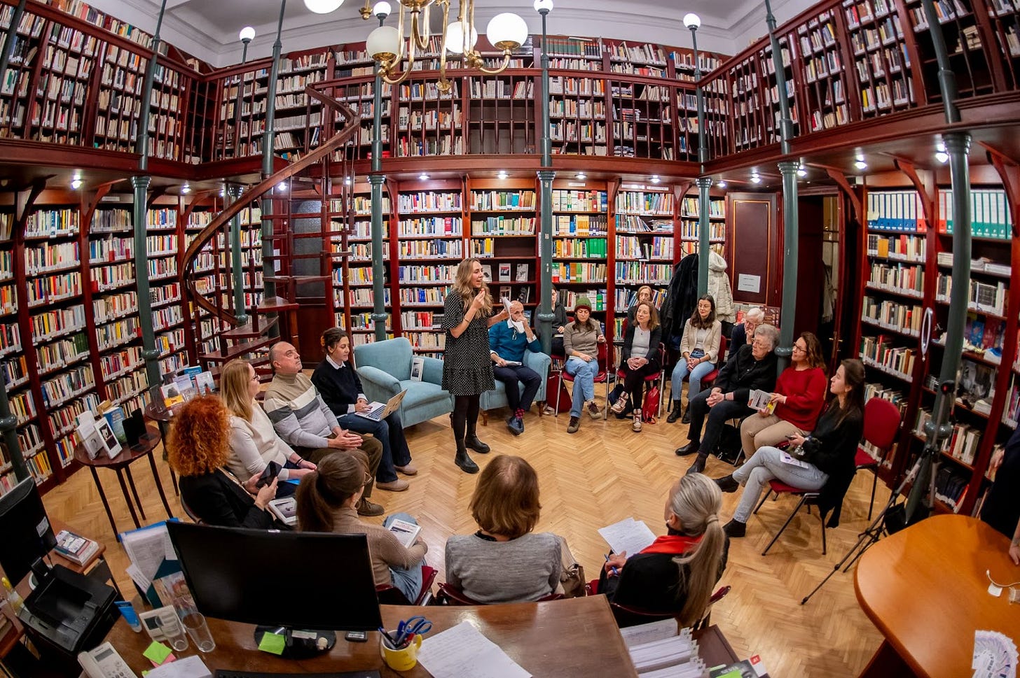 Un incontro del Club del Libro nella Biblioteca dell'Istituto Italiano di Cultura di Budapest; la foto ritrae una sala con pareti interamente coperte da scaffali di libri posti su due piani; c'è una scala di legno ricurva che porta da un piano all'altro e un lampadario a grandi bulbi bianchi; la sala è attrezzata con poltrone e sedie, ci sono diversi partecipanti al Club e, al centro, Sveva Borla. e