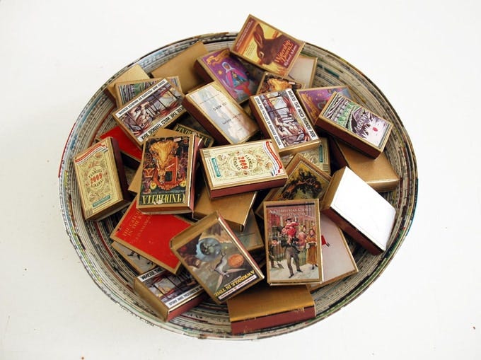 A round, shallow woven basket filled with miniature matchbox-style books. Each tiny book has a colorful, vintage-inspired cover design featuring various illustrations and typography, such as animals, historical scenes, and ornate lettering. The boxes appear to be handmade or artistically crafted, resembling collectible items or novelty mini books. A round, shallow woven basket filled with miniature matchbox-style books. Each tiny book has a colorful, vintage-inspired cover design featuring various illustrations and typography, such as animals, historical scenes, and ornate lettering. The boxes appear to be handmade or artistically crafted, resembling collectible items or novelty mini books.