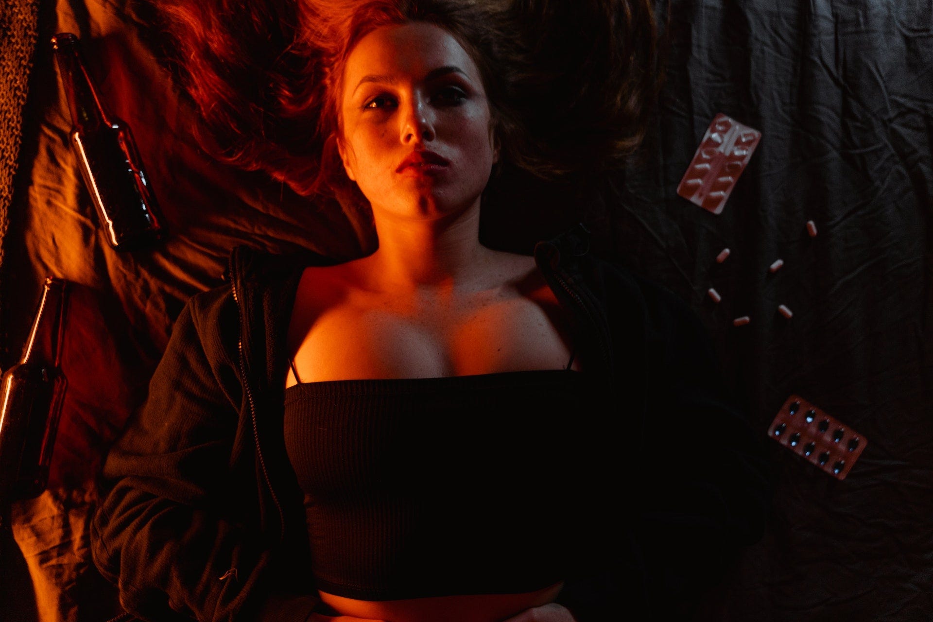 A woman on a bed in the dark with bottles and pills around her. A woman on a bed in the dark with bottles and pills around her.