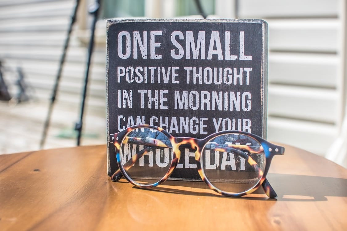 A wooden sign with the message "One small positive thought in the morning can change your whole day" and a pair of glasses in front. A wooden sign with the message "One small positive thought in the morning can change your whole day" and a pair of glasses in front.